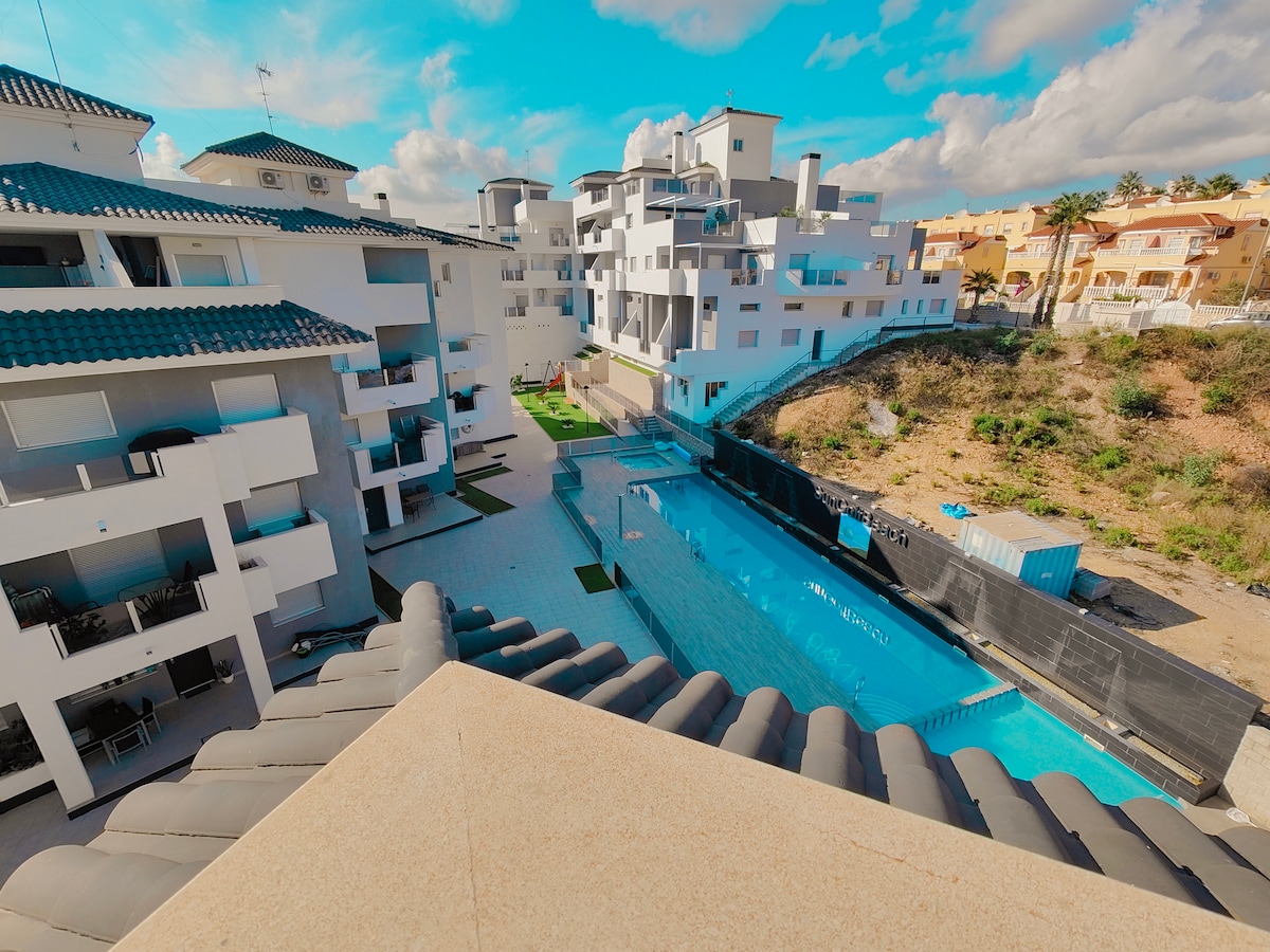 An overview of the community area is showcased, featuring a large blue swimming pool surrounded by landscaped greenery. The modern apartment buildings are positioned around the pool, with balconies offering a view of the outdoor space. A clear sky and scattered clouds provide a bright backdrop.