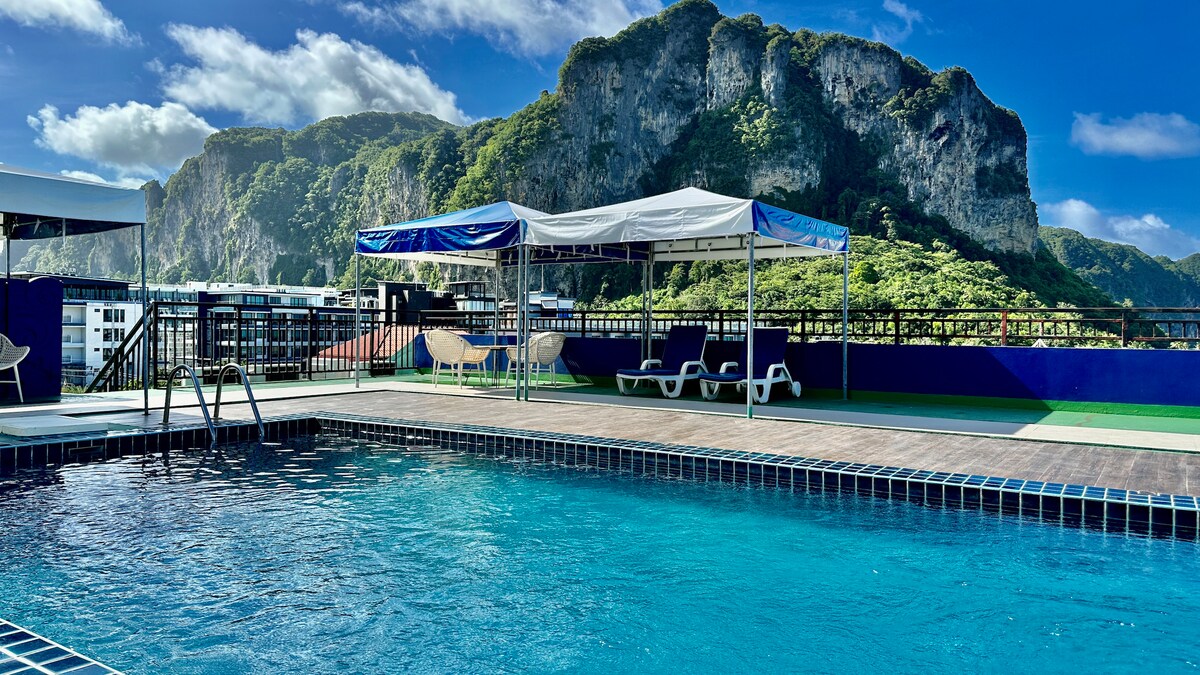 A rooftop swimming pool is visible, featuring clear blue water and a wooden deck. Two shaded areas with white canopies provide seating options, while a scenic view of verdant hills is displayed in the background, enhancing the tranquil environment.