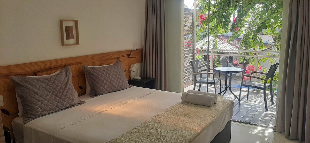 A comfortable sleeping area is complemented by large windows that open to a serene outdoor space. A spacious bed adorned with soft pillows sits next to a nightstand. A small table and chairs can be seen on the balcony, surrounded by lush greenery.