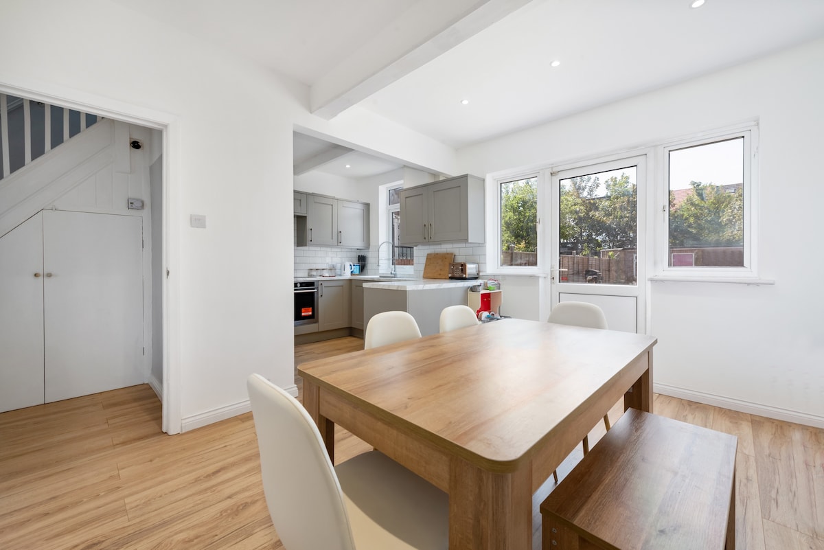 An open plan dining area is showcased, featuring a wooden table surrounded by white chairs. Natural light filters through large windows, illuminating the space. The adjoining kitchen displays modern cabinetry and appliances, with a staircase visible in the background.