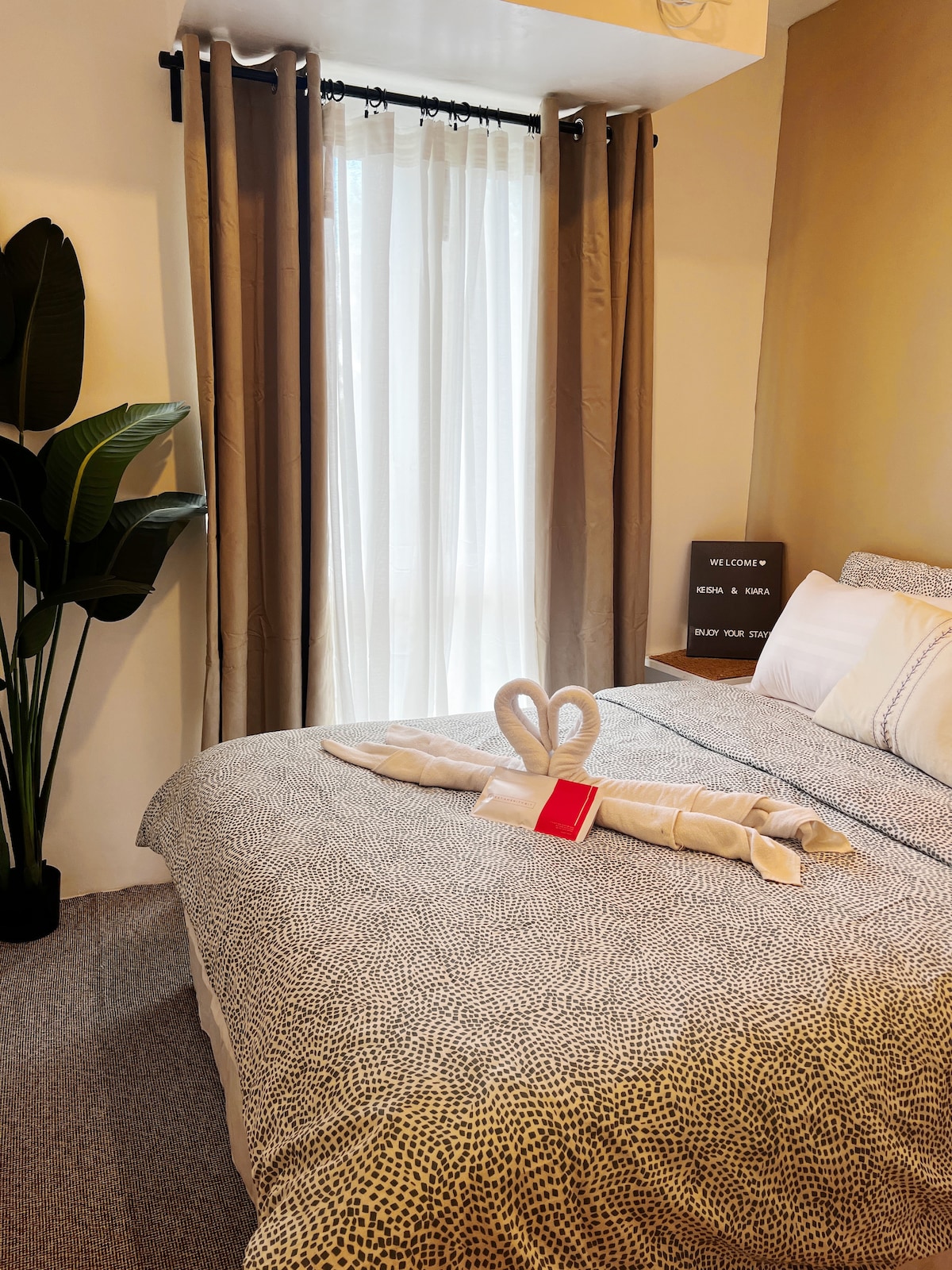 A relaxing bedroom features a spacious bed adorned with patterned bedding and neatly arranged pillows. Natural light filters through sheer curtains, highlighting a potted plant in the corner. A welcome sign sits on a side table, creating an inviting atmosphere for guests.