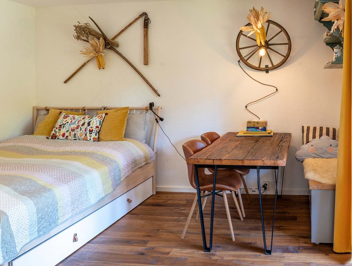 A cozy sleeping area features a double bed dressed in a patterned quilt, accompanied by decorative pillows. A wooden table with stylish chairs sits in the corner, complemented by wall decor made of natural materials. Soft lighting creates an inviting space.