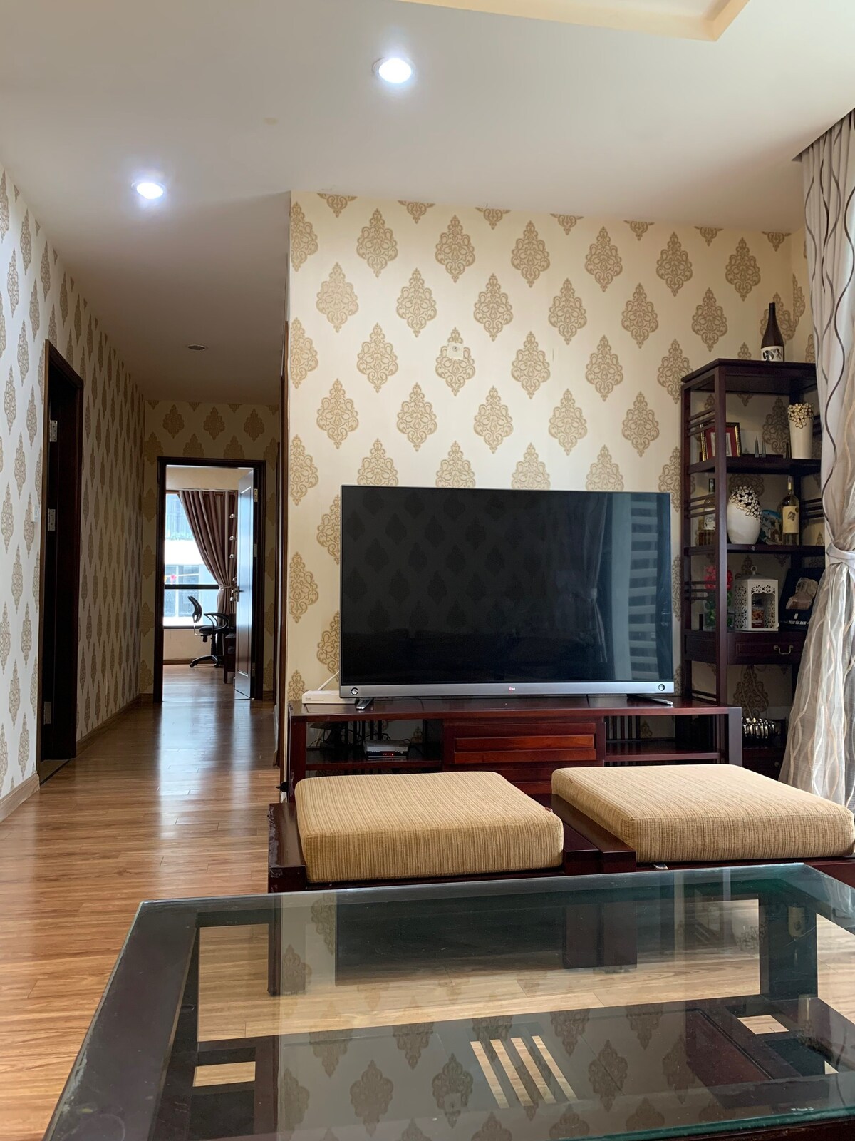 An inviting living area features a large flat-screen television positioned on a wooden console. A glass coffee table sits in front, accompanied by two cushioned ottomans. The patterned wallpaper adds visual interest, while a doorway in the background leads to another room with a view.