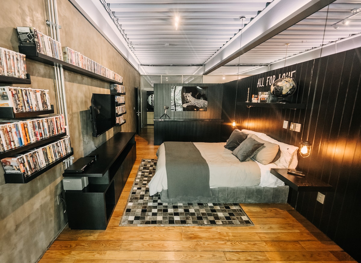 A modern bedroom is showcased with a spacious bed centered on a wooden floor. Movie shelves line one wall, featuring a variety of DVDs. A sleek TV is mounted across from the bed, while ambient lighting is provided by stylish pendant lamps.