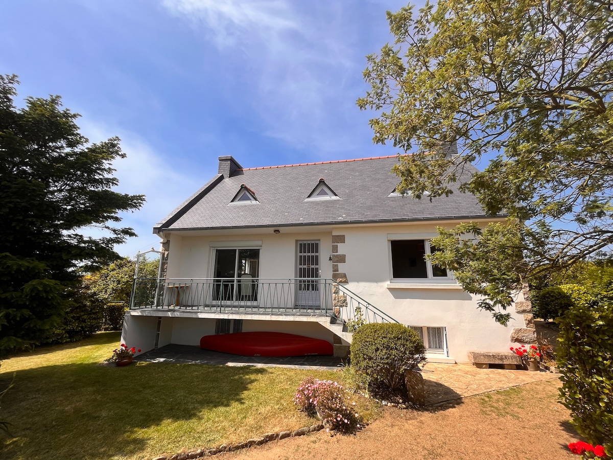 A spacious house features a welcoming exterior with a sloped roof and a broad balcony. Surrounding greenery includes trees and shrubs, with a neatly maintained lawn in the foreground. A bright red canoe is placed against the house, enhancing the outdoor space.