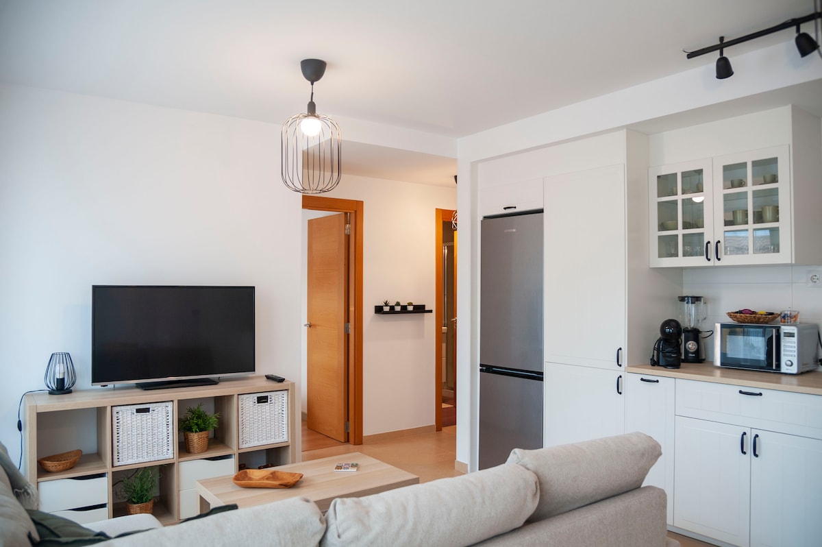 A well-lit living area features a comfortable sofa and a coffee table. A television is positioned on a media unit with storage baskets. The kitchen area is equipped with modern appliances, including a refrigerator, microwave, and coffee maker, all set within a clean and organized layout.