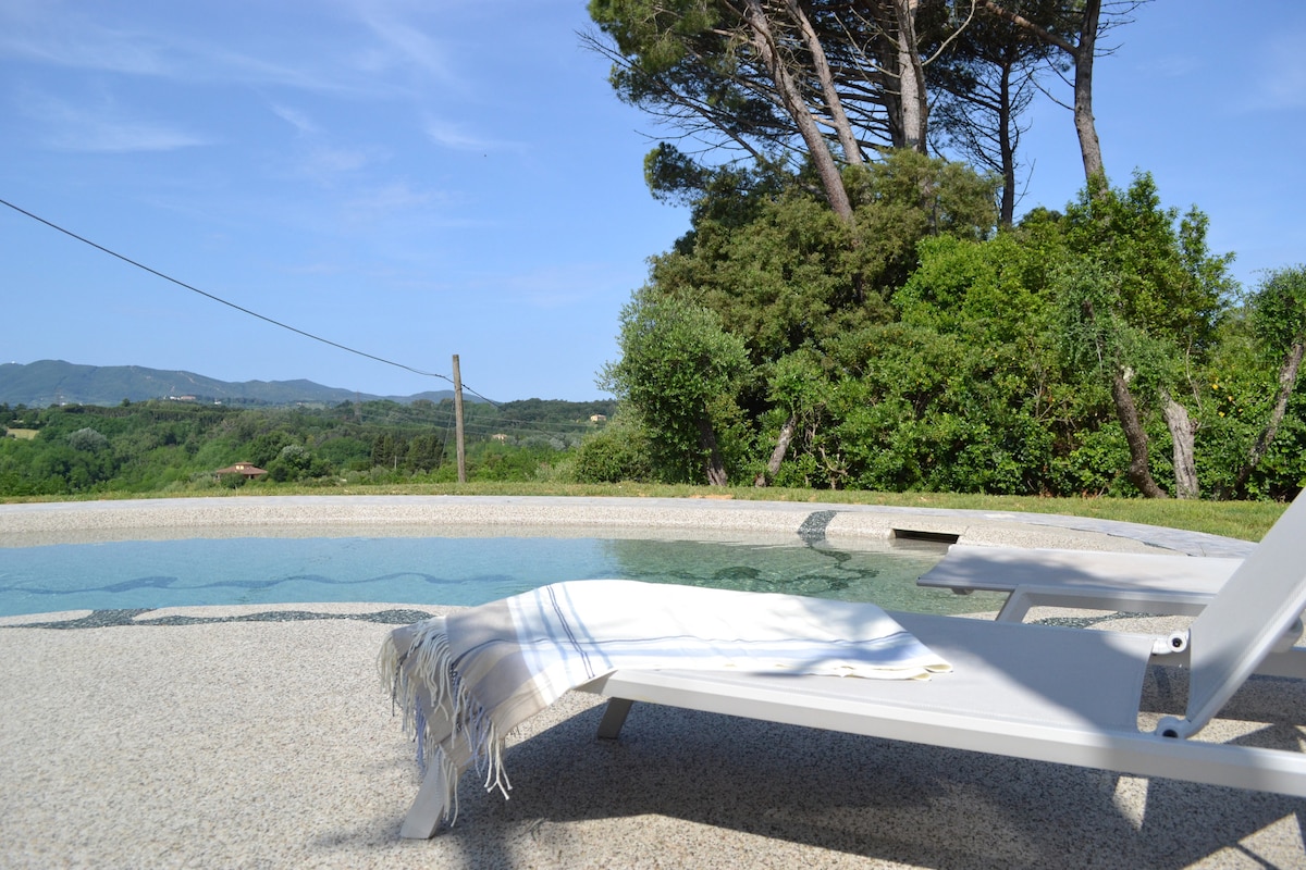 A sunlounger is positioned beside a unique pool, featuring hydrotherapy features. Surrounding greenery and hills are visible, with a clear blue sky above, creating a serene outdoor atmosphere. A white towel is laid across the lounger, inviting relaxation.