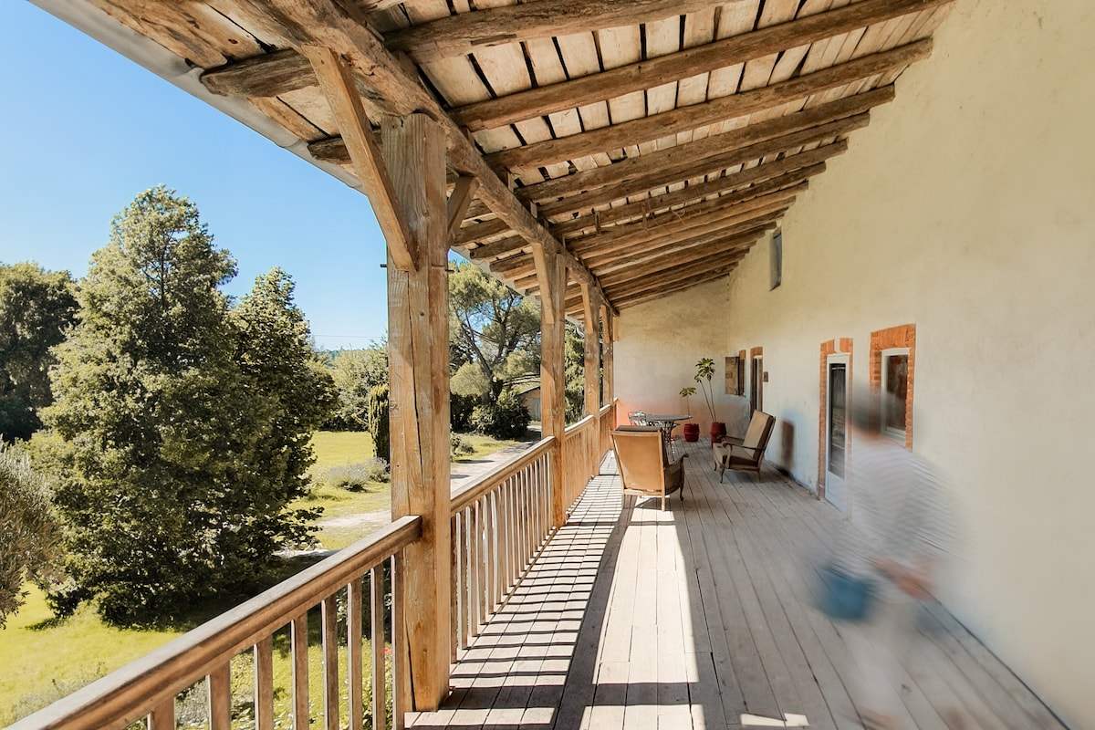 A spacious wooden terrace features a rustic design with exposed beams and a long balcony railing. Two comfortable chairs are arranged for relaxation, overlooking lush greenery. Natural light fills the area, enhancing the inviting outdoor space that connects with the surrounding landscape.