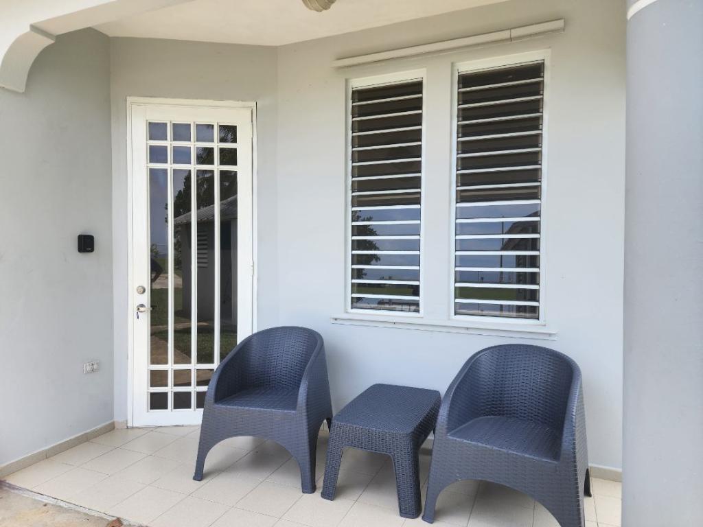 A small, inviting outdoor seating area features two blue wicker chairs and a matching side table. Natural light fills the space through white-shuttered windows, allowing views of the outside. A glass-paned door provides access to the interior.