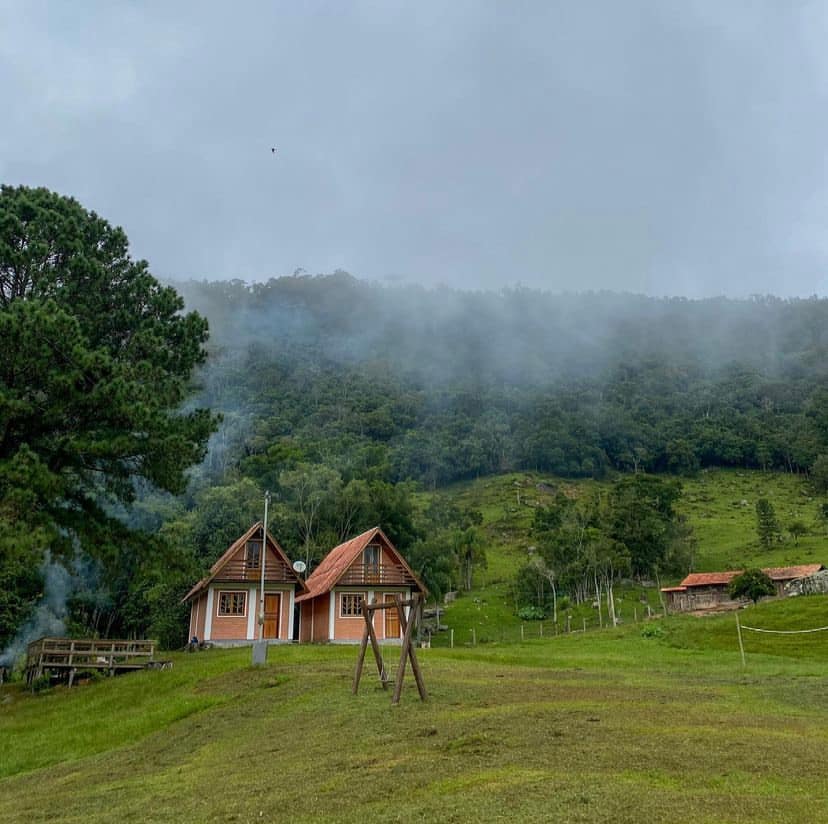 The charming cabin is set amidst lush green hills, surrounded by mist. A swing set is visible in the foreground, while the cabin features a sloped roof and wooden accents. Trees and mountain scenery complete the natural landscape, providing a serene atmosphere.