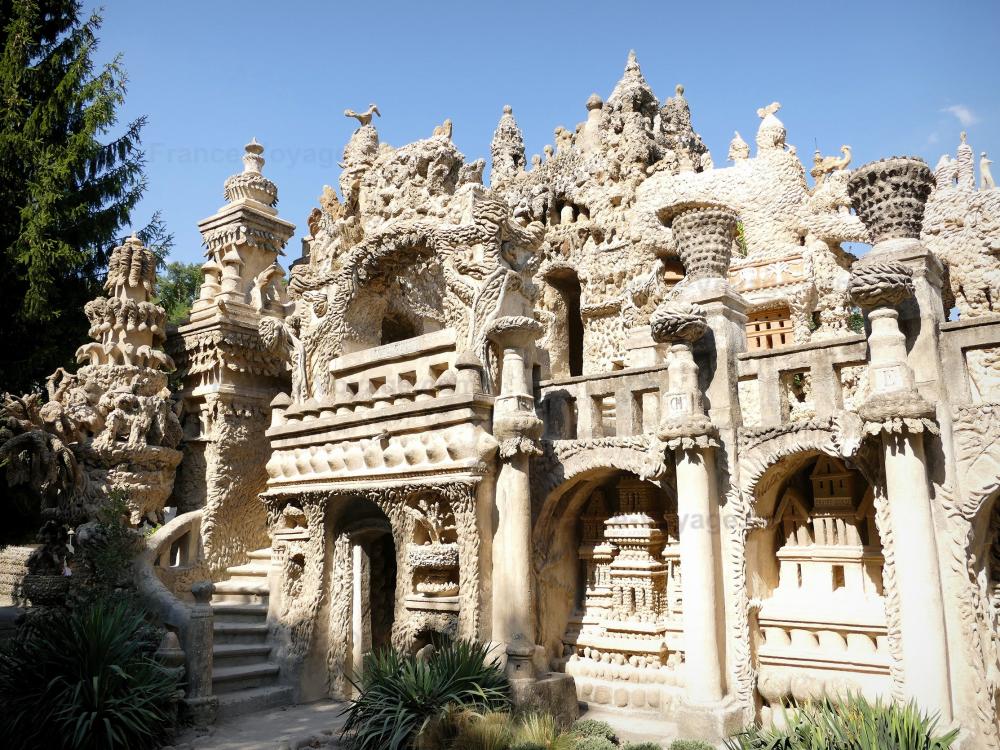 An intricate stone structure showcases elaborate carvings and sculptures, depicting various ornamental designs. The façade features a combination of arches, columns, and tiers with foliage surrounding the building, under a clear blue sky. The surroundings are adorned with greenery, enhancing the artistic details.