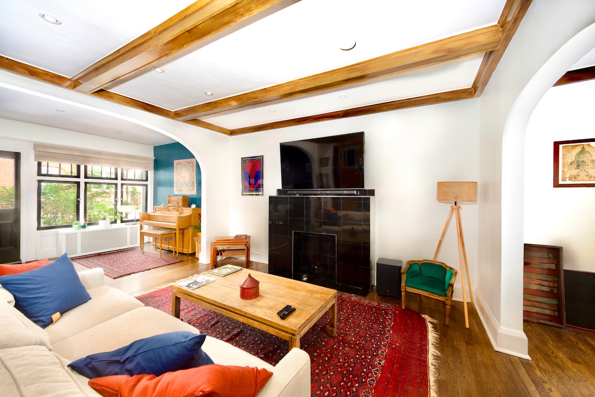 The spacious living area features a comfortable sofa adorned with colorful throw pillows. A large television is mounted on a sleek black unit, with a wooden coffee table positioned centrally on a decorative rug. Natural light floods in from multiple windows, highlighting the room's elegant design.