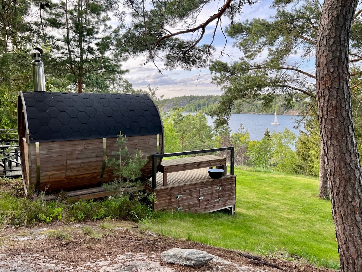 A wooden sauna, shaped like a barrel, is set within a green landscape, surrounded by trees. A small deck area with a bowl sits beside the sauna, offering views of the nearby water and distant sailboat, enhancing the tranquil outdoor setting.