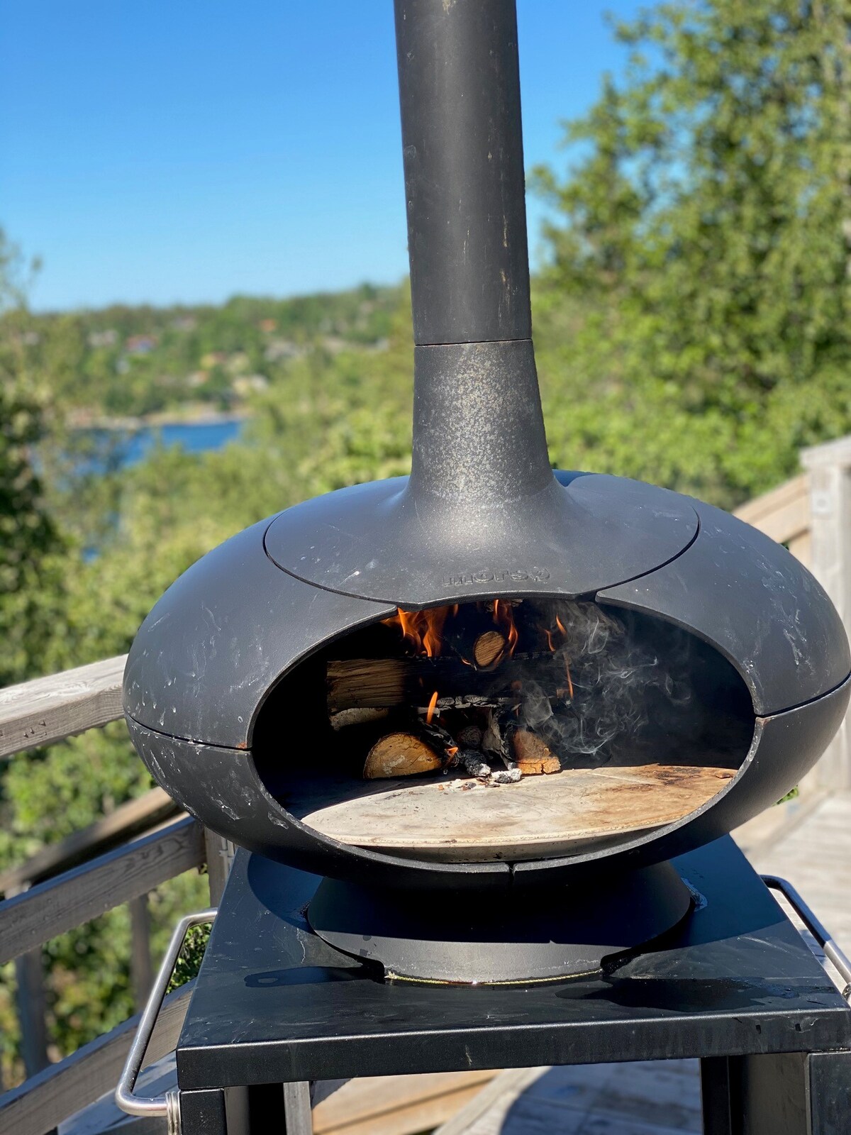 An outdoor pizza oven is positioned on a deck, showcasing a wood-fired interior with flames visible. Surrounding greenery and a distant view of water are captured in the background, emphasizing a serene outdoor setting.