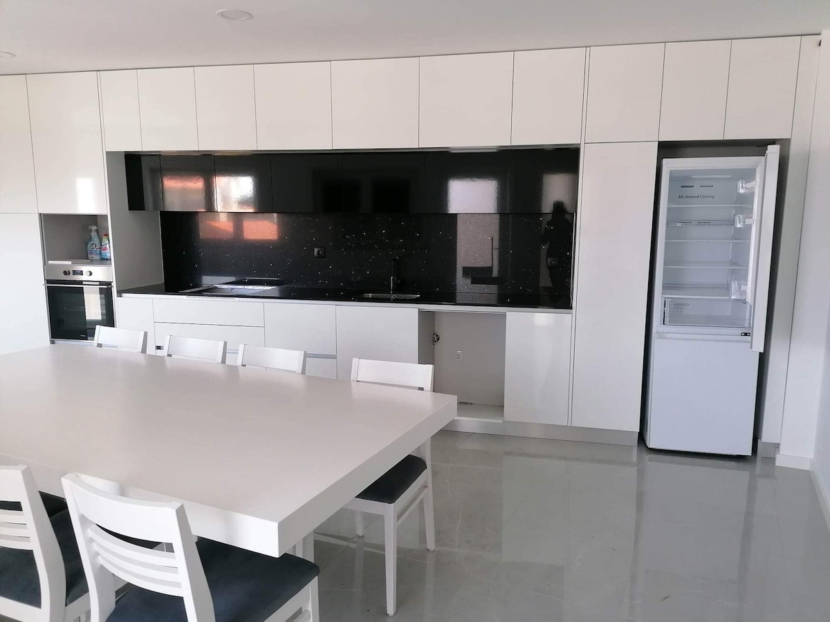An elegant kitchen is presented, featuring sleek white cabinetry with black countertops. A spacious dining table is set with white chairs, and modern appliances, including an oven and refrigerator, are visible. Natural light filters through a window, enhancing the functional design.