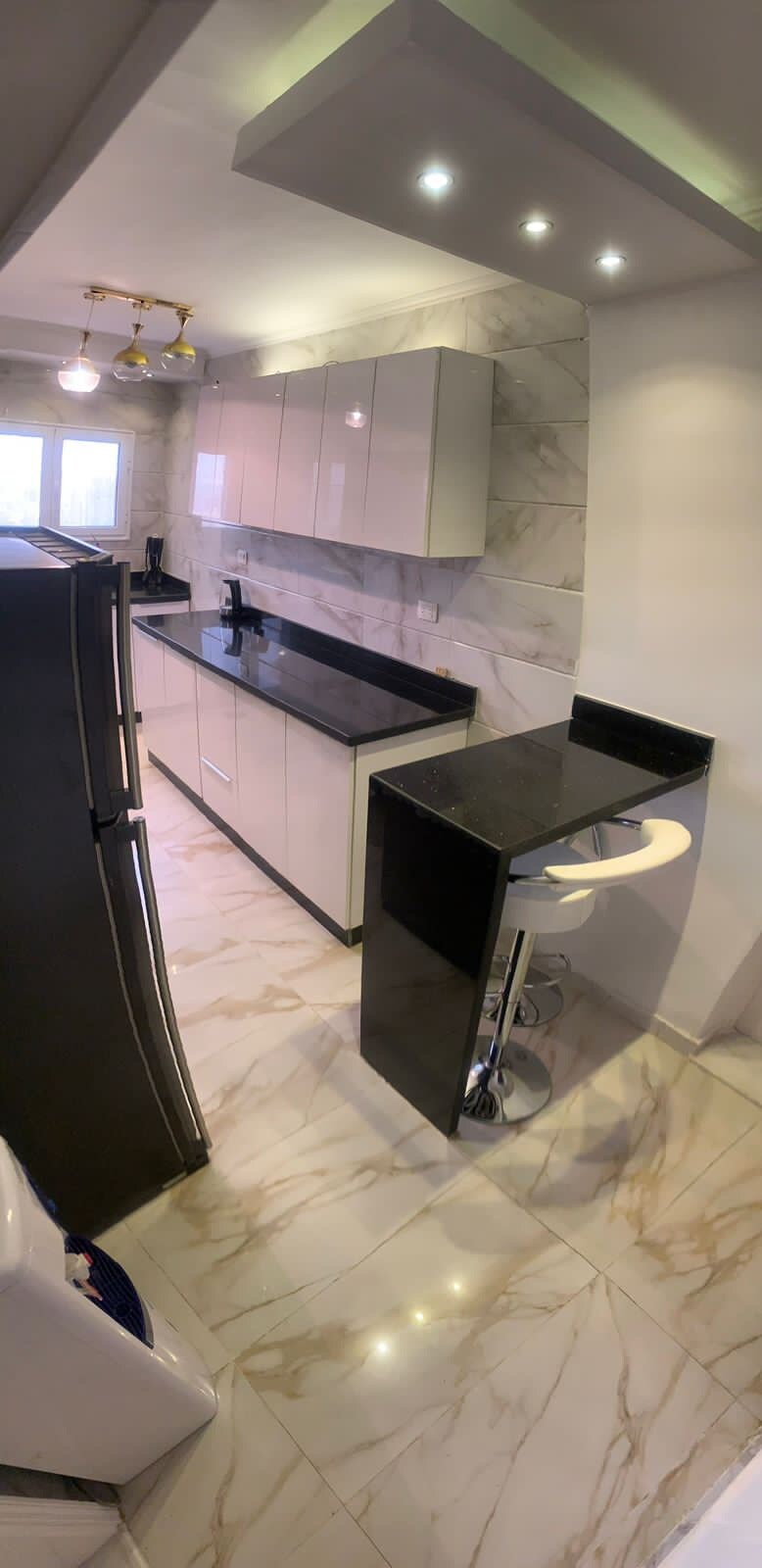 The kitchen features sleek cabinet storage and a black countertop that contrasts with white cabinetry. A modern barstool is positioned at a compact counter space, complemented by warm lighting fixtures above. A marble-patterned floor adds an elegant touch to the area.