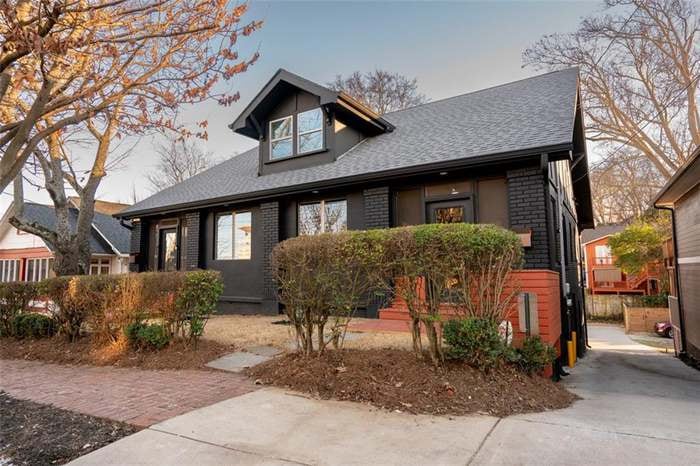 The exterior of a charming 1920s home is presented, featuring a black-painted façade with large front windows. Lush green bushes line the front yard, while a pathway leads to the entrance. The well-maintained lawn adds to the inviting appearance of the property.