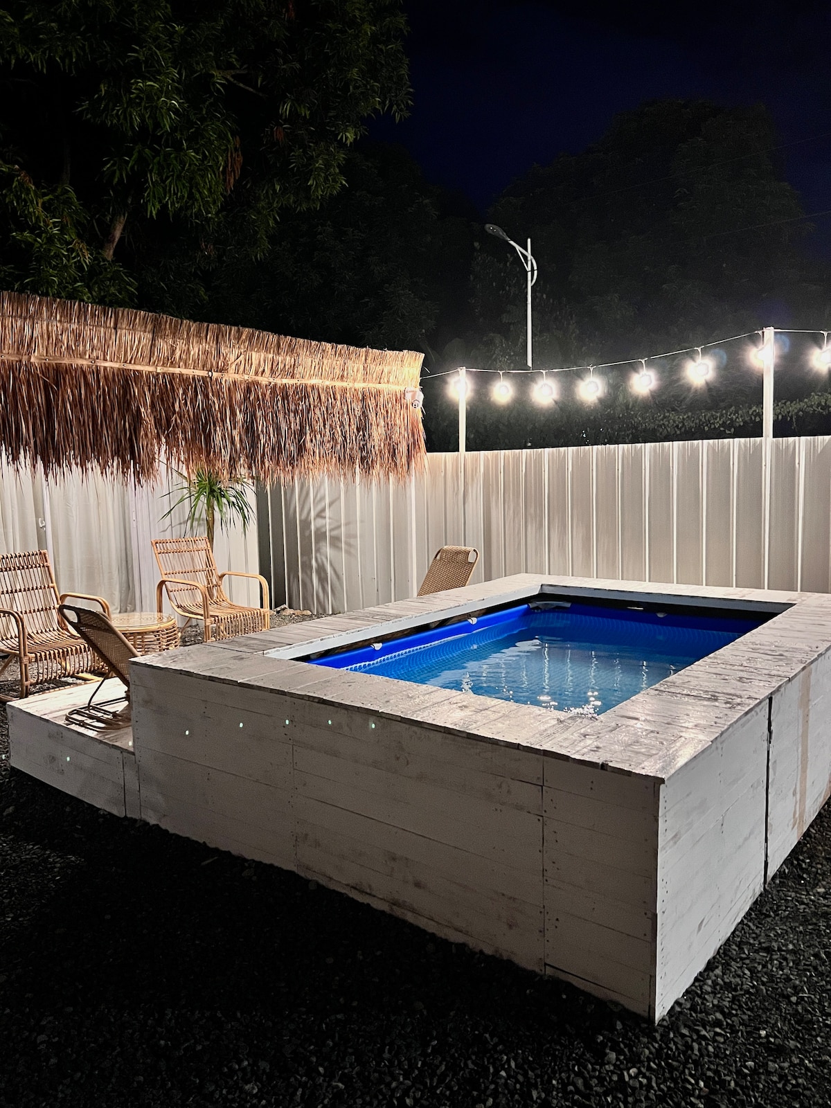 A plunge pool is surrounded by a wooden deck, creating a cozy outdoor area. Soft string lights overhead illuminate the space, enhancing the inviting atmosphere. Two chairs are positioned nearby, with tropical foliage providing additional greenery.