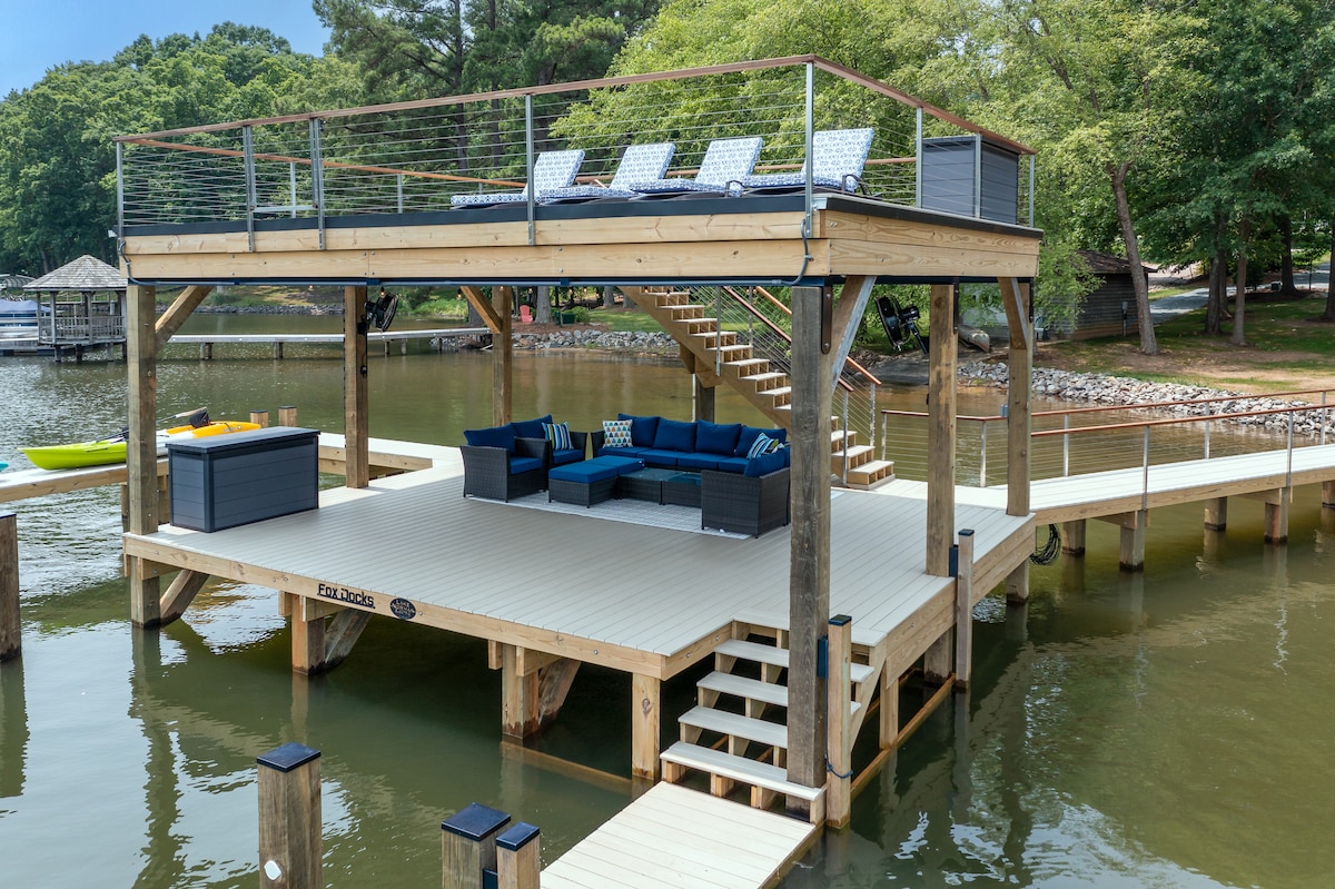 A two-level dock is presented, featuring a spacious sundeck with lounge chairs and a comfortable outdoor seating area below. Surrounded by serene waters, the structure invites relaxation and leisure activities, with kayaks visible in the background.