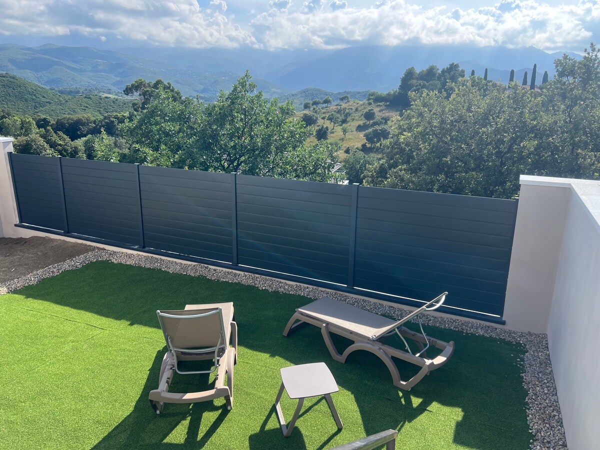 A private terrace features two lounge chairs and a small side table, surrounded by artificial grass. A panoramic view of rolling hills and greenery is visible beyond a modern fence, with soft, natural lighting enhancing the serene outdoor space.
