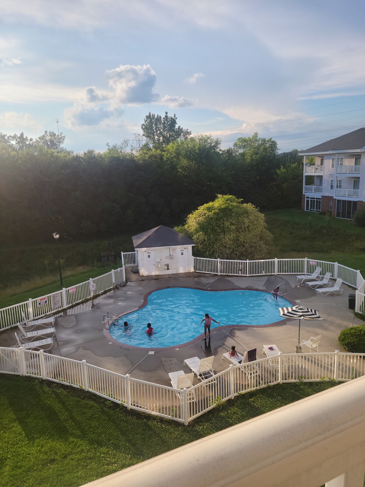 A pool area is surrounded by a white fence and includes a small gazebo. The clear water reflects the sky, and several lounge chairs are positioned around the pool. Green trees provide a natural backdrop, creating a serene environment.