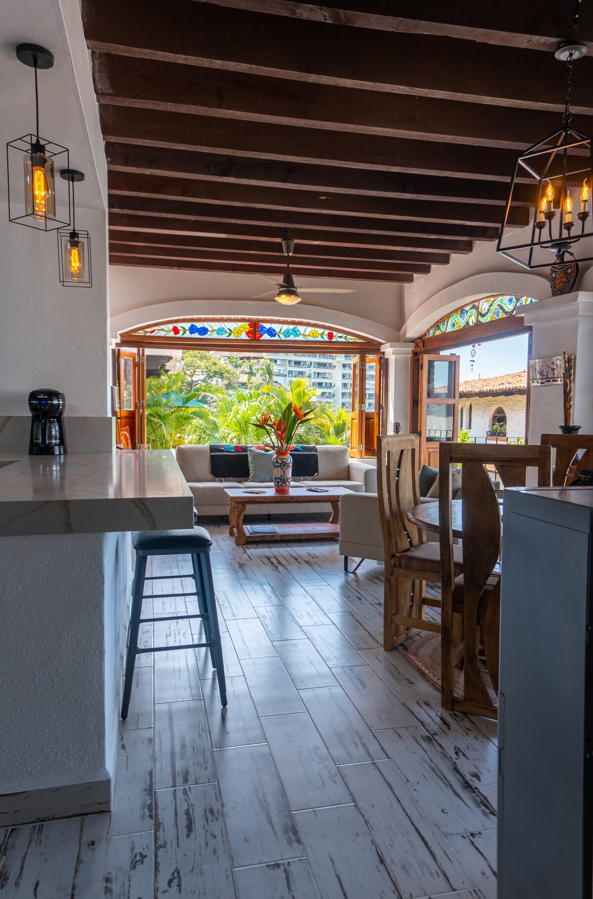 A bright open living area is characterized by high wooden ceilings and arched stained glass windows that frame views of lush greenery. A cozy seating arrangement is complemented by a wooden dining table, and modern light fixtures hang above, enhancing the vibrant atmosphere.