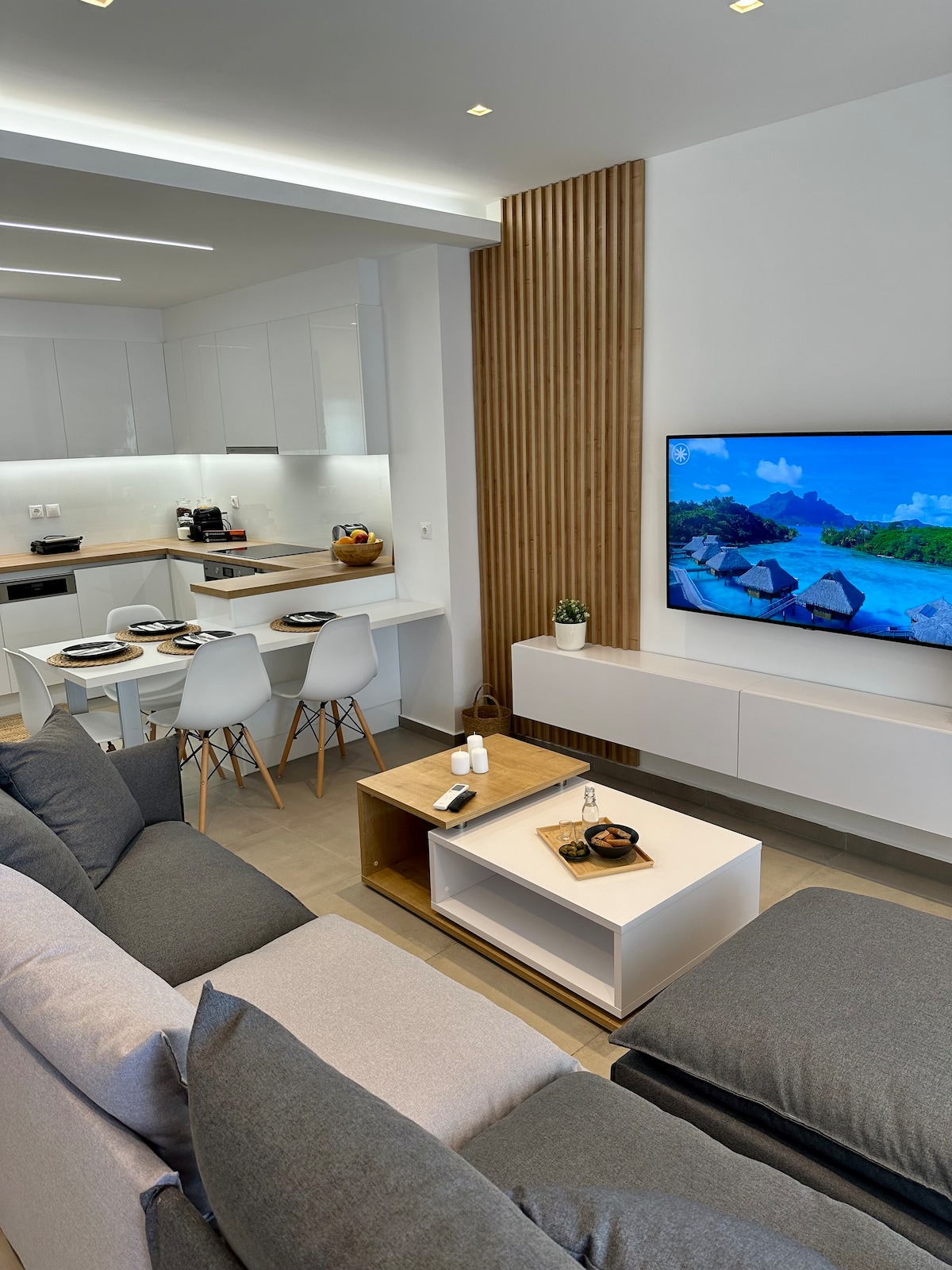 An open living area is presented, showcasing a comfortable grey sofa facing a wall-mounted television. A light wood coffee table holds decorative items. In the background, a dining area features a round table set with plates and sleek white chairs, accompanied by contemporary kitchen elements.