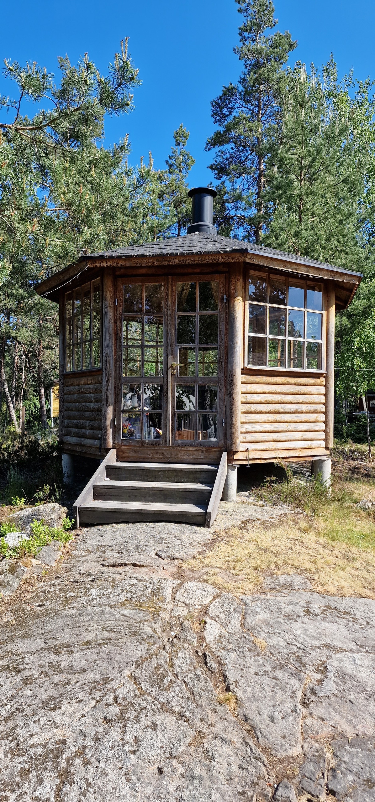 A rustic grill house is depicted, featuring a circular wooden structure with large windows. The entry is accessed by a set of stairs leading up from a natural stone surface. Surrounding pine trees offer a cozy and serene backdrop.