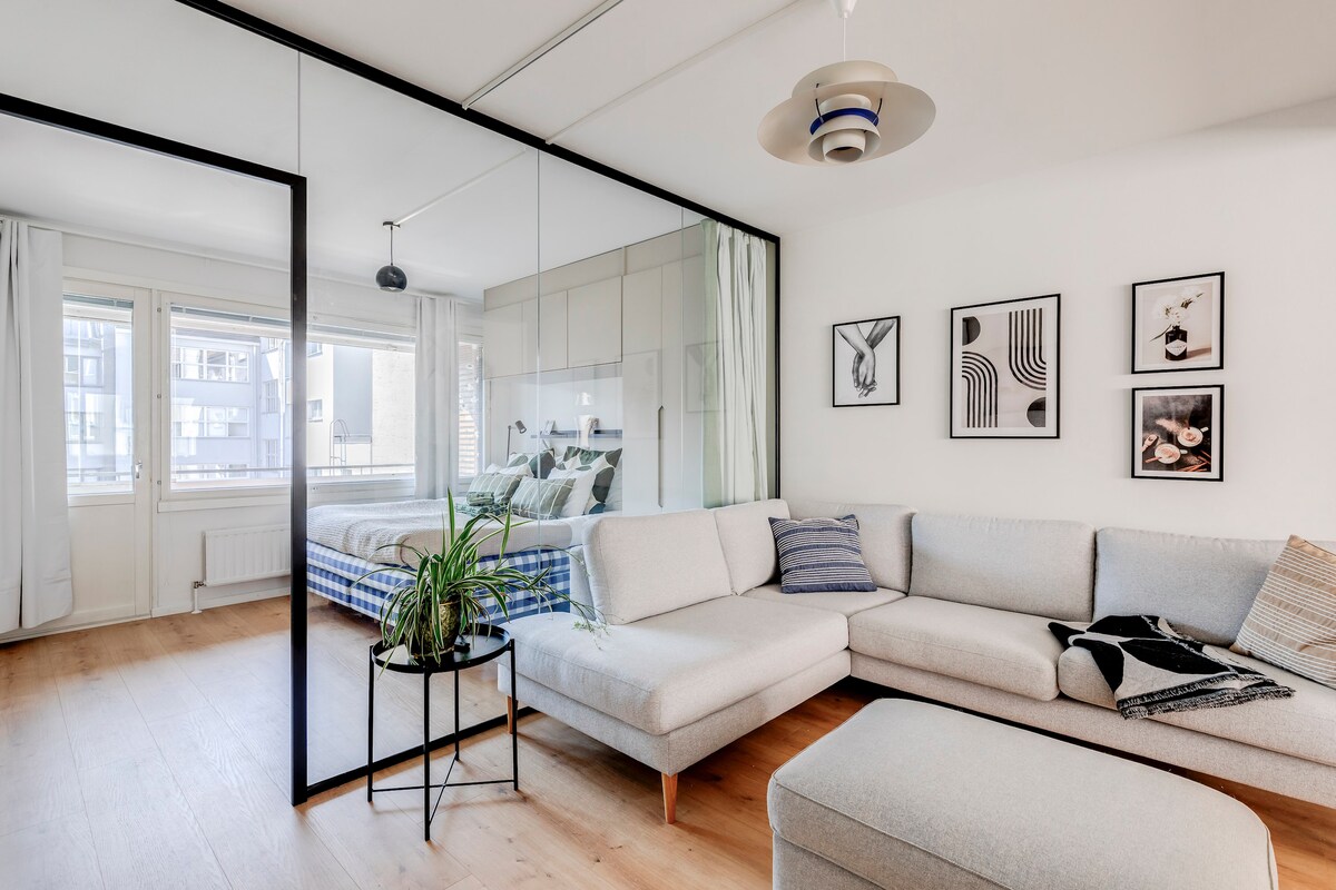 A modern living space is showcased, featuring a light-colored sectional sofa positioned near a glass partition. The sleeping area is visible beyond, with a spacious bed and large windows allowing natural light. Framed art pieces adorn the walls, contributing to the contemporary aesthetic.