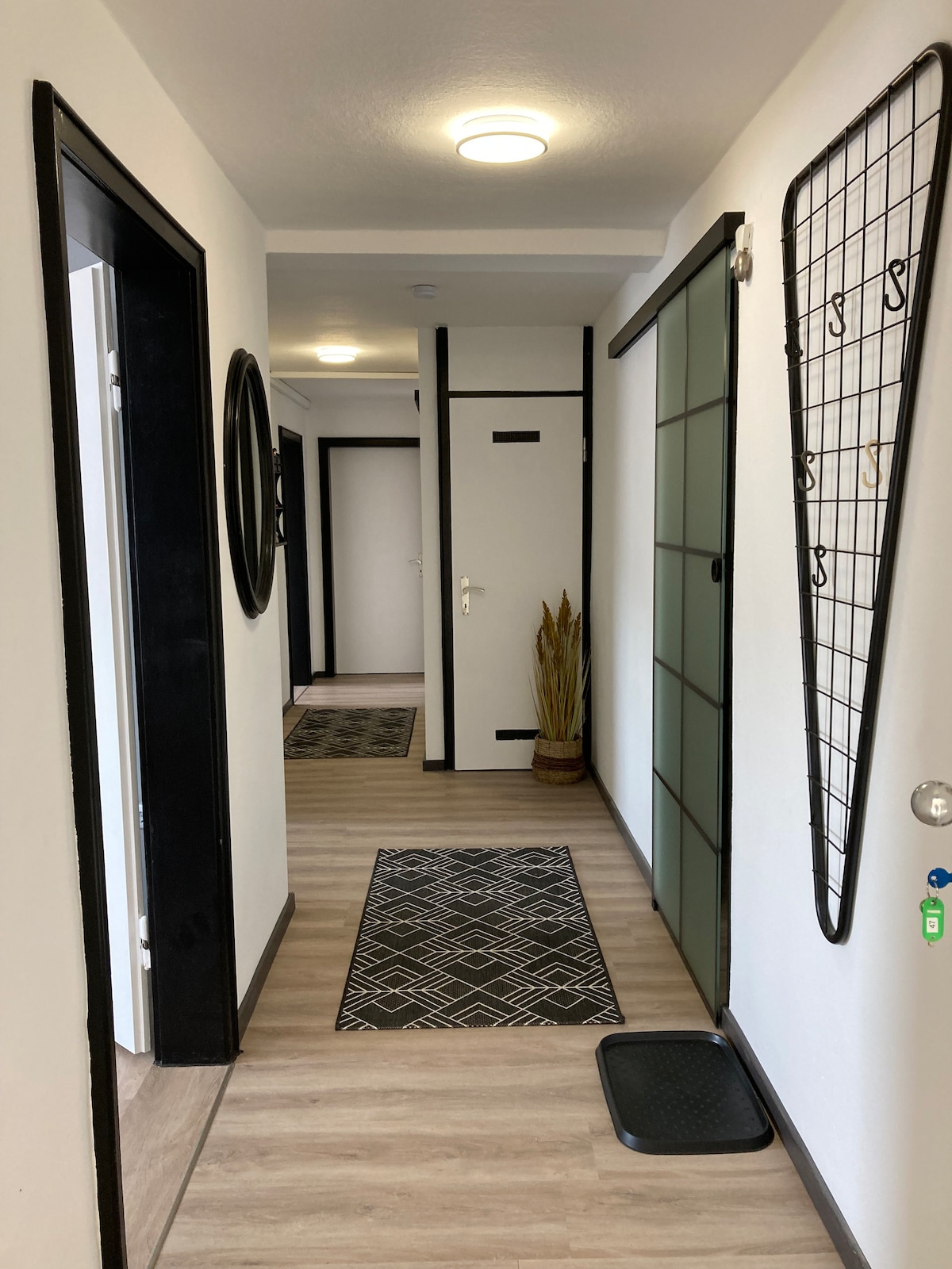 A well-lit hallway features light wood flooring and white walls, creating a spacious feel. Decorative elements include a round mirror and a floor plant. Two black-framed doors lead to rooms, and geometric-patterned rugs add warmth to the entrance area.