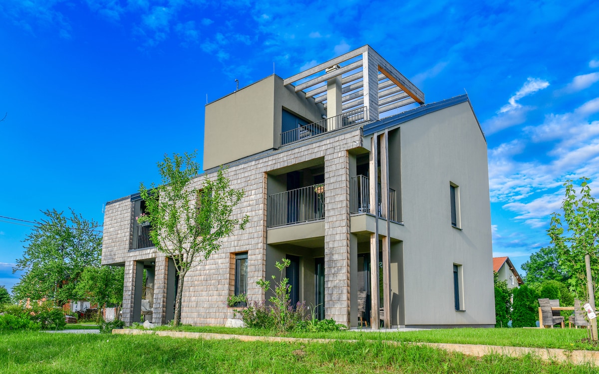 A modern Lika house is depicted, featuring a combination of stone and sleek gray facades. Balconies are visible on the upper levels, while landscaped greenery surrounds the structure. The sky is bright and clear, complementing the contemporary design.