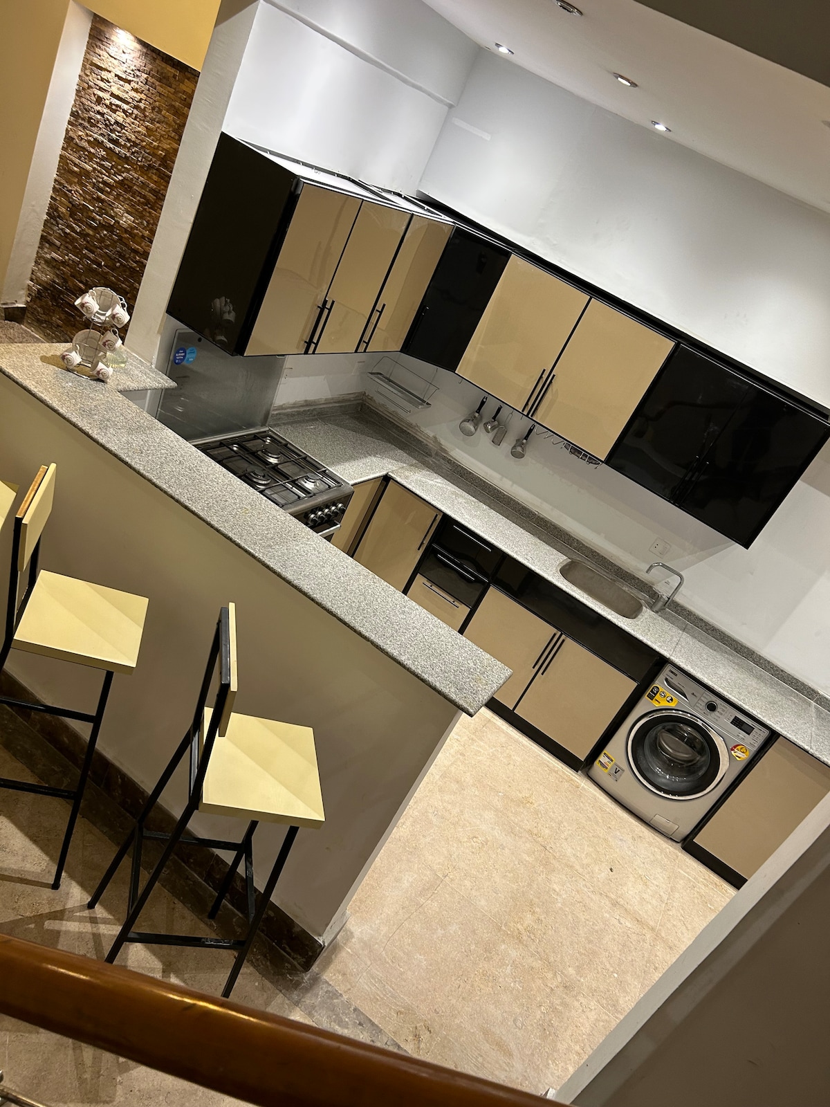 The kitchen features modern cabinetry in a two-tone design, complemented by a gray countertop. A stove, washing machine, and various kitchen appliances are positioned for convenience. Two high-backed chairs are situated at the counter, enhancing functionality in the space.