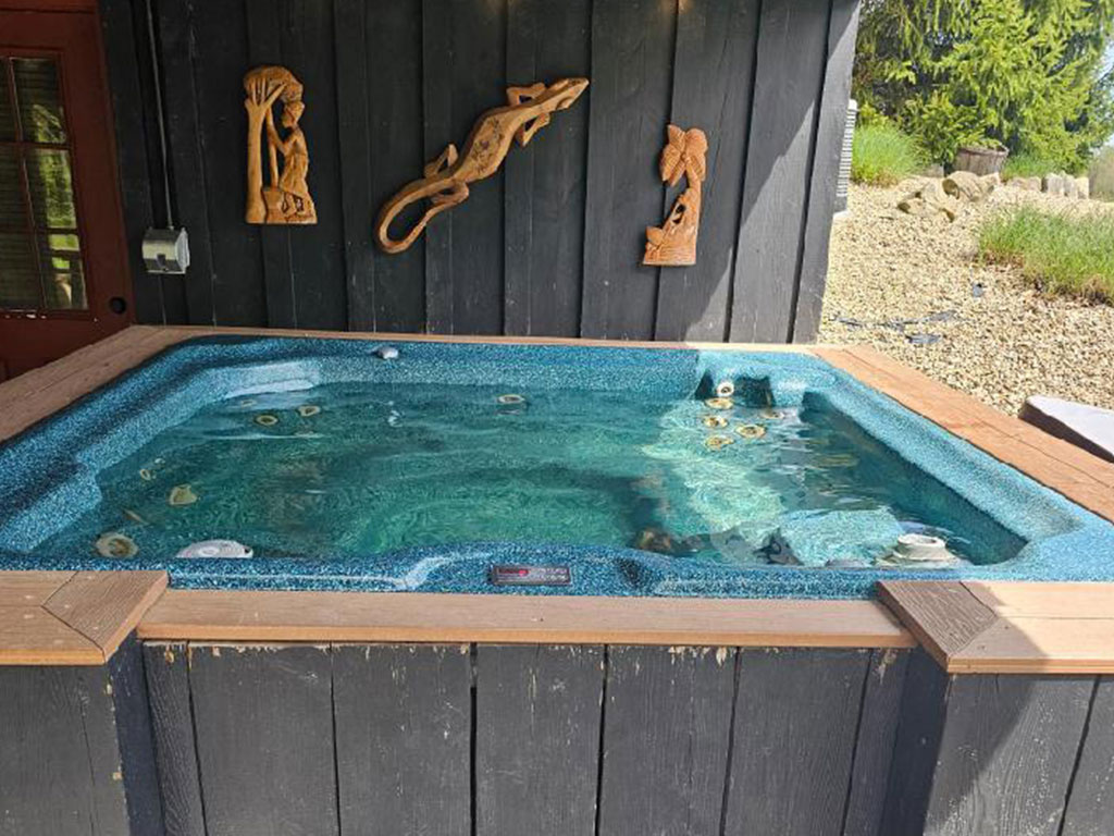 A private hot tub is nestled on a wooden deck, surrounded by a natural setting. The water shimmers, reflecting light as wooden accents, including art pieces, adorn the nearby walls. Relaxation opportunities are prominently featured in this tranquil outdoor space.