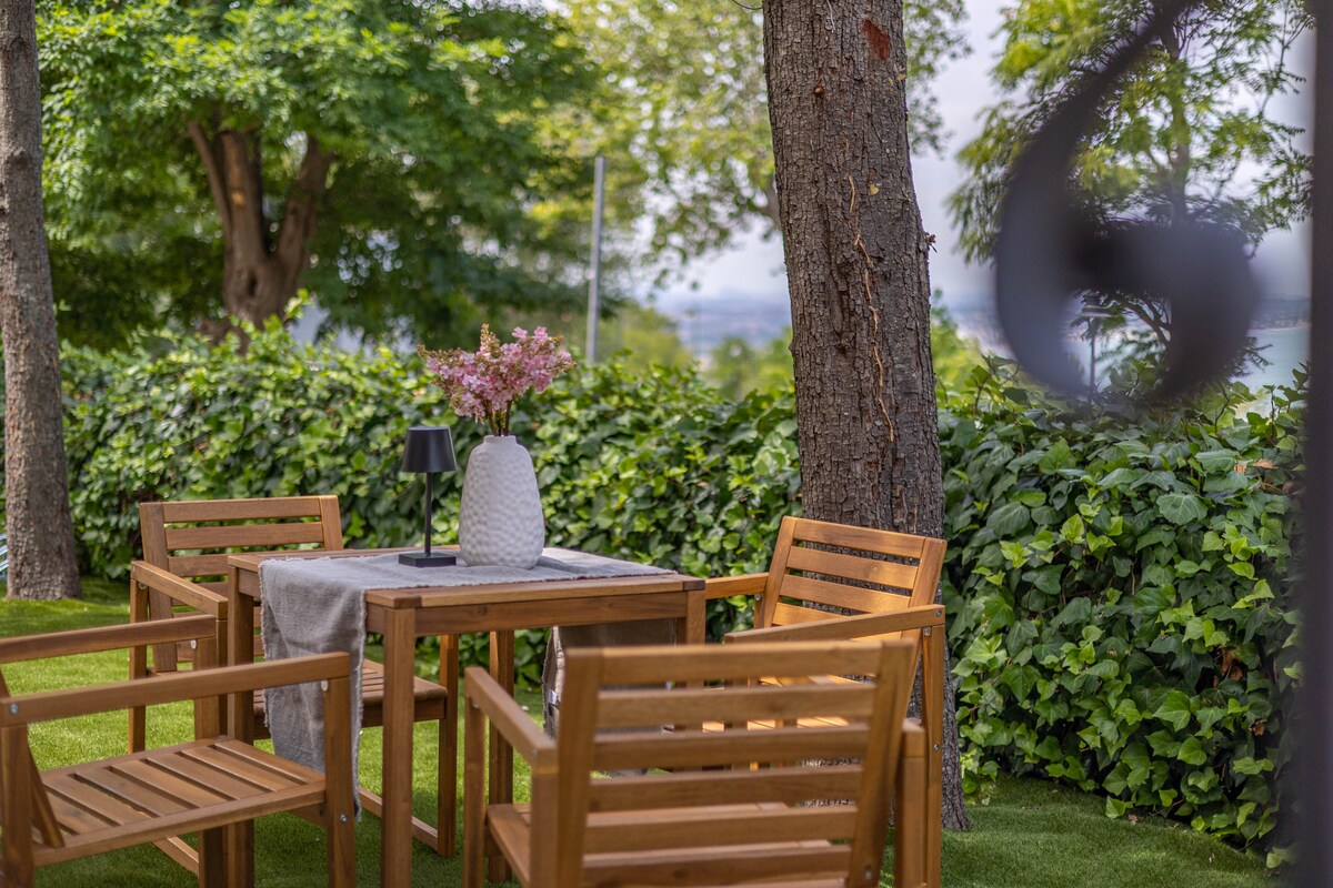 A shared outdoor garden area features a wooden table surrounded by four matching chairs. A vase with flowers decorates the table, and the space is bordered by lush greenery and tall trees, providing a serene setting for relaxation.