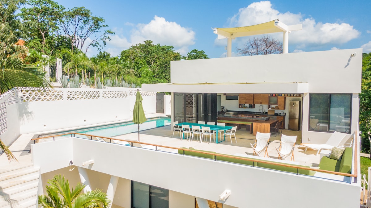 The image showcases a modern two-story Casa featuring a large outdoor terrace with a private pool. The seamless integration of indoor and outdoor spaces is highlighted by expansive glass doors, while lush greenery surrounds the property, providing a serene atmosphere.
