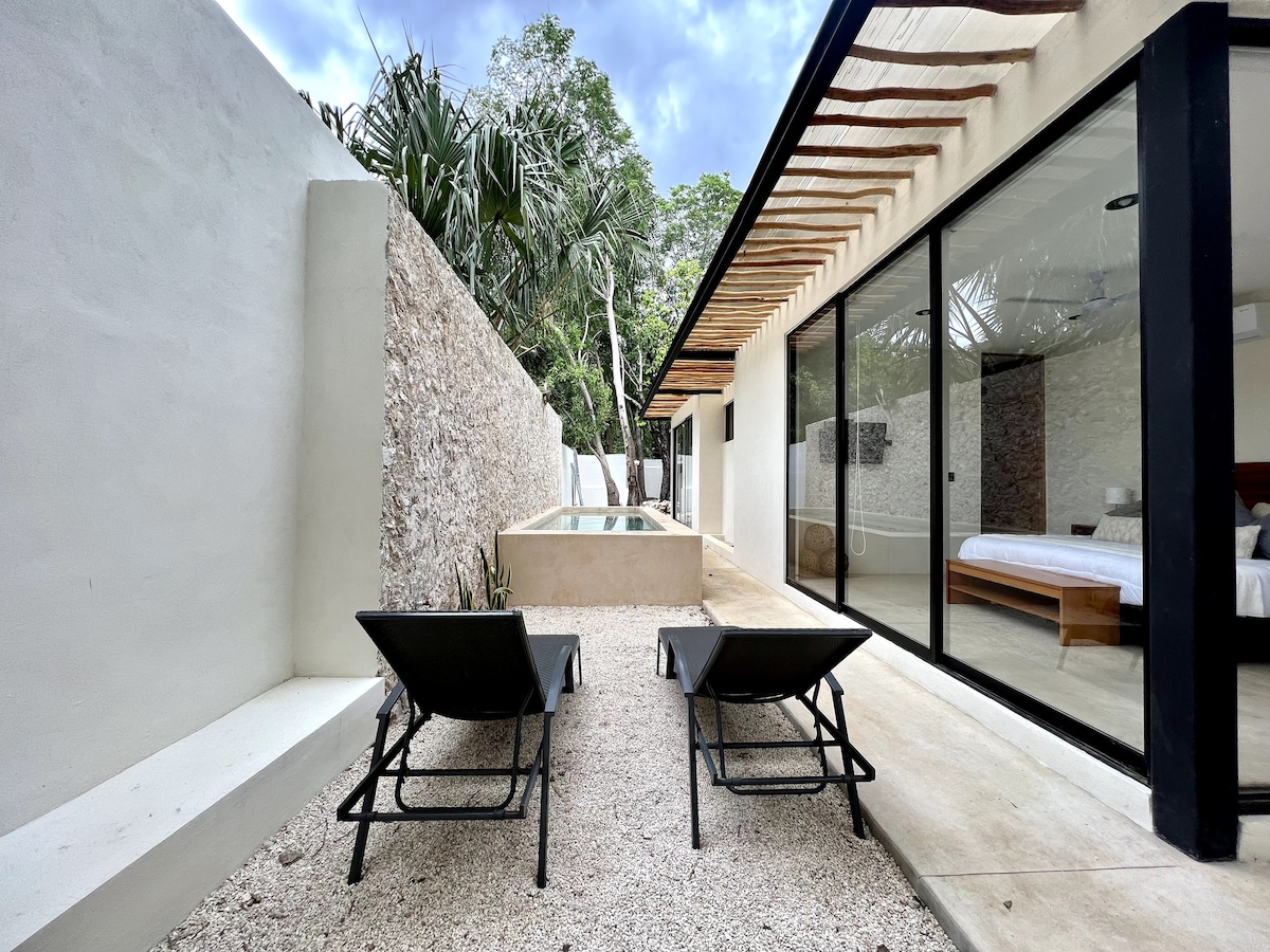 A serene outdoor space features two lounge chairs positioned near a small pool. The area is surrounded by a stone wall and lush greenery, with large glass doors leading to an interior sleeping space filled with natural light.