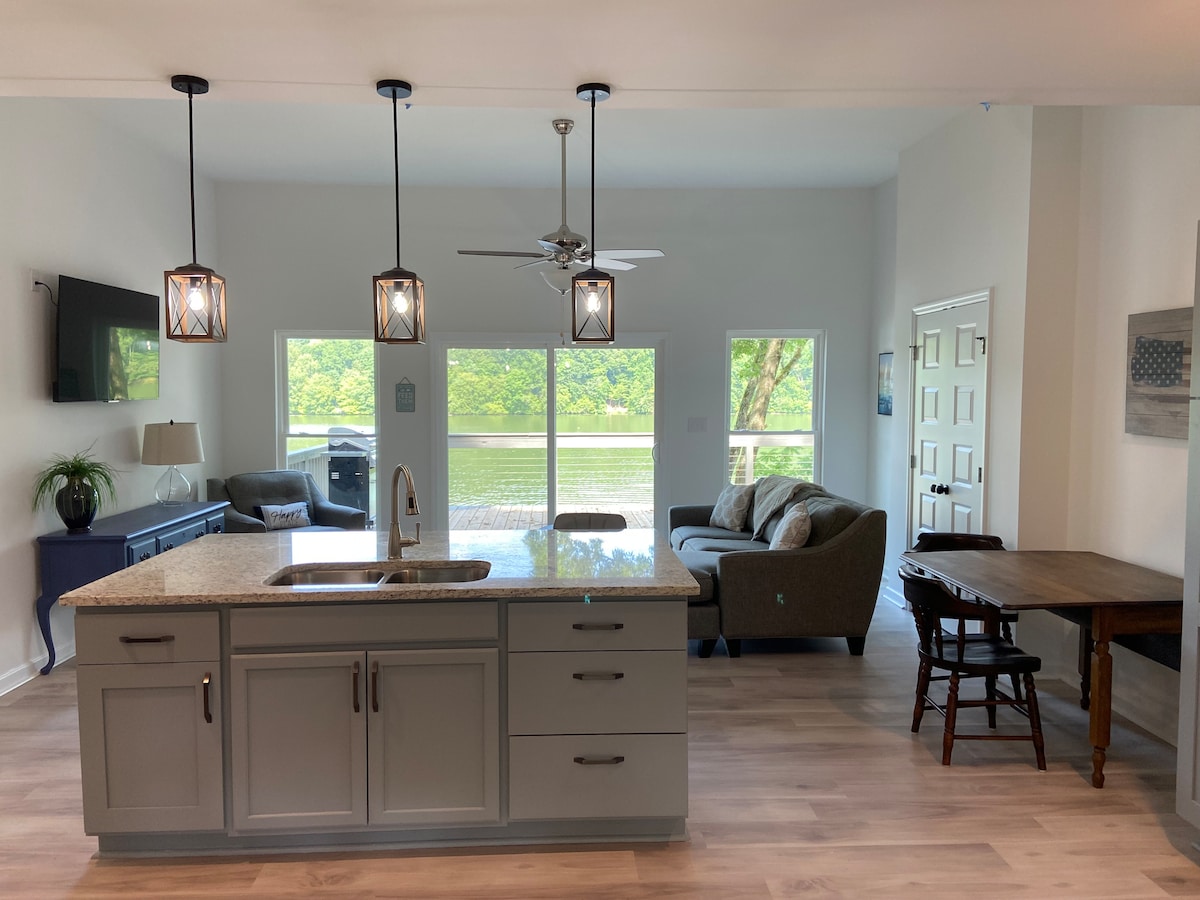 The modern kitchen features a spacious countertop with a sink and pendant lighting above. An open concept design allows views of the living area and large windows that showcase serene river scenery. A dining table and comfortable seating are present, inviting relaxation.
