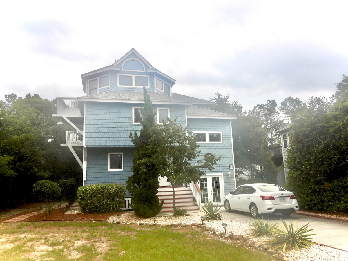 The three-story beach house, painted in shades of blue, features multiple decks and a wide staircase leading to the entrance. Lush greenery surrounds the property, enhancing the welcoming exterior. A white vehicle is parked in the driveway, adding to the home-like atmosphere.