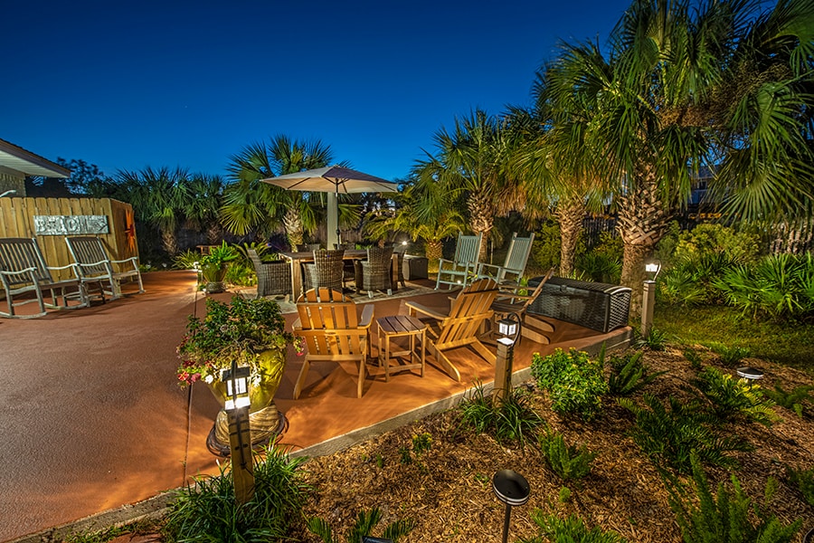 A spacious outdoor patio is highlighted by comfortable seating arrangements under a large umbrella. Soft landscape lighting illuminates the area, accentuating the surrounding palm trees and vibrant plant life, creating a serene atmosphere for relaxation in the evening.
