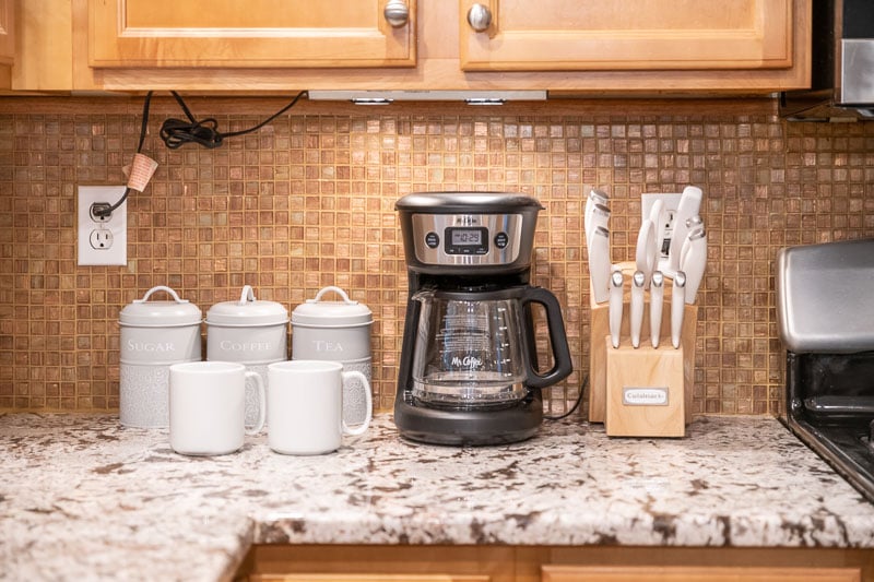 A kitchen counter is arranged with a coffee maker, two white mugs, and a set of ceramic canisters labeled for sugar, coffee, and tea. A wooden knife block displays five knives. The backdrop features a textured mosaic tile wall.