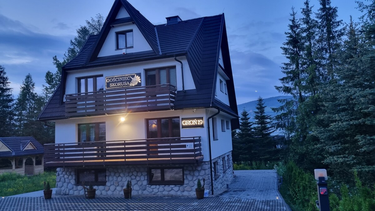 A multi-story building with a sloped roof and stone foundation is shown at dusk. The exterior features wooden balconies on the upper levels. Surrounding greenery includes tall trees and a paved parking area in the foreground.