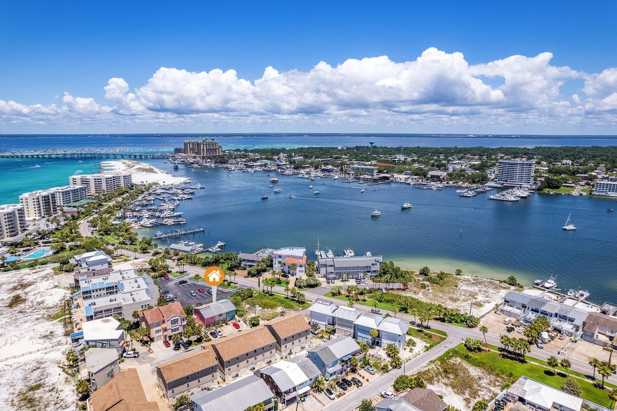 An aerial view captures the vibrant coastal landscape of Destin, showcasing a blend of marina, white sandy beaches, and the azure Gulf waters. Nearby, various boats are anchored, and a mix of buildings line the shore, reflecting the lively atmosphere of the area.