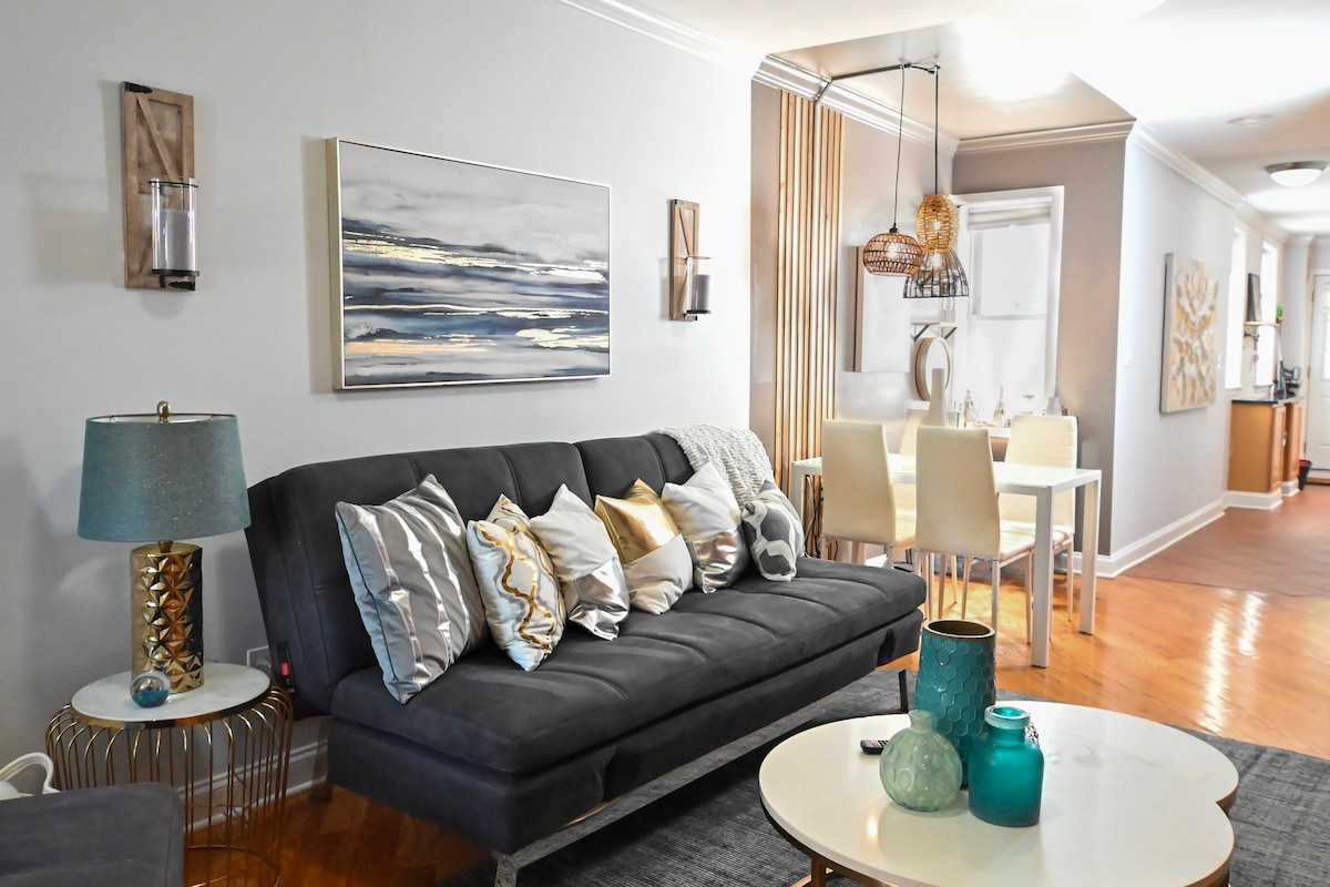 The living area features a comfortable couch adorned with decorative pillows. A round coffee table sits in front, complemented by a side table with a lamp. A dining area with a light-colored table and chairs is visible in the background, enhancing the open floor plan.