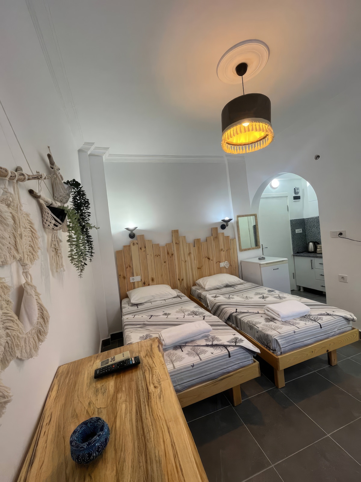 Two individual beds with light-patterned linens are positioned against a wooden accent wall. A wooden table sits nearby with decorative items. The space is illuminated by a pendant light and features a kitchenette visible through an archway.