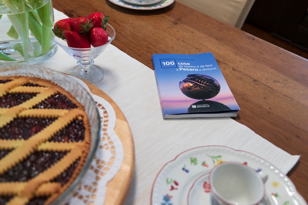 A wooden dining table is set with a decorative lace tablecloth. A glass bowl of strawberries is adjacent to a book titled '100 cose da vedere e da fare a Pesaro e dintorni.' A pie, appearing freshly baked, rests on a delicate serving platter.