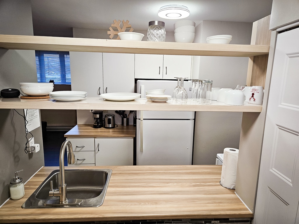 A modern kitchen showcases open shelving with neatly arranged dishes and glassware. A stainless steel sink is positioned in front of a polished countertop, with a coffee maker and refrigerator visible in the background. Natural light enters through a window, illuminating the space.