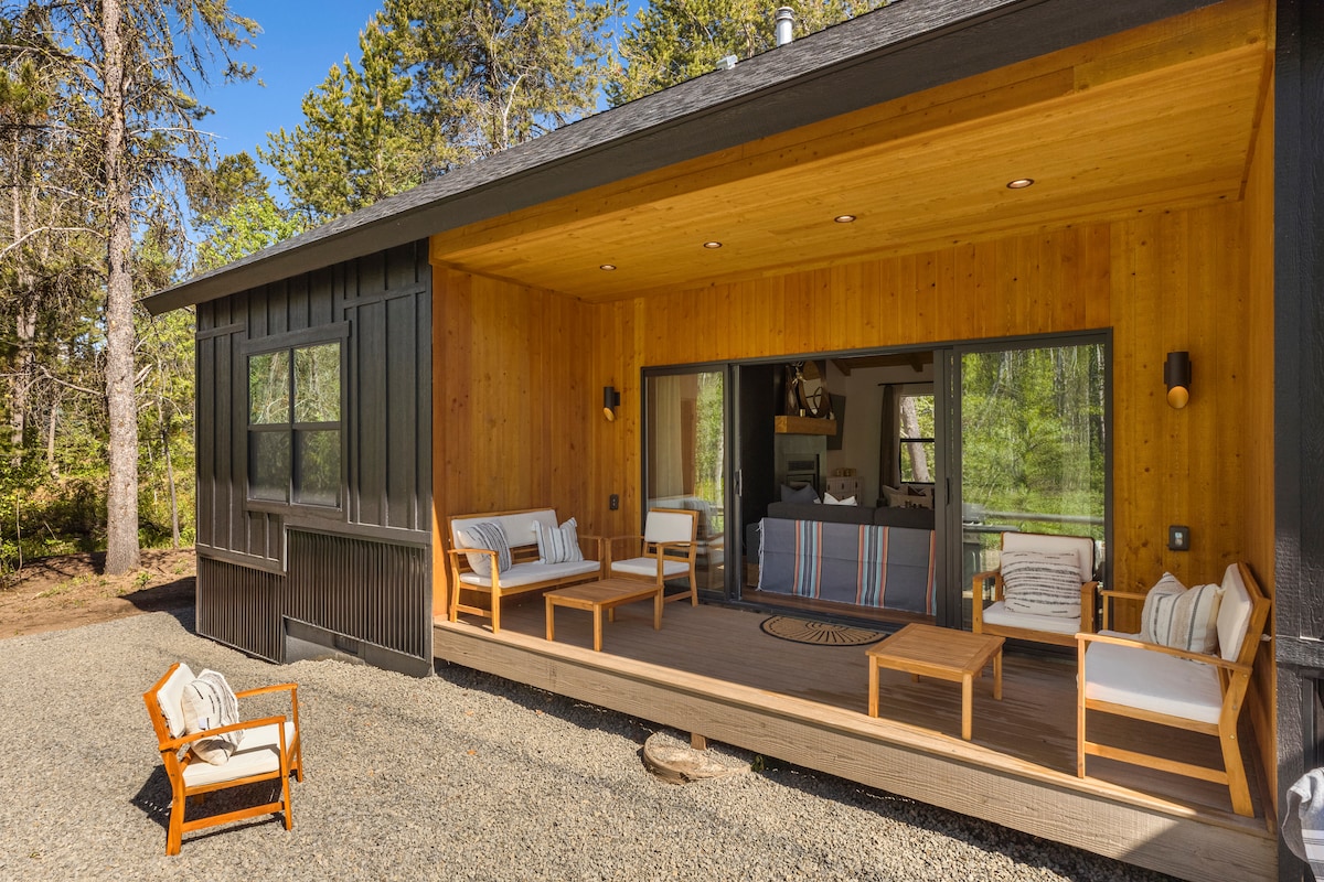 An outdoor living space features wooden flooring and a covered porch area, providing comfortable seating with light-colored cushions. Large sliding glass doors offer a view into the interior, while surrounding trees add a natural backdrop to the relaxing setting.
