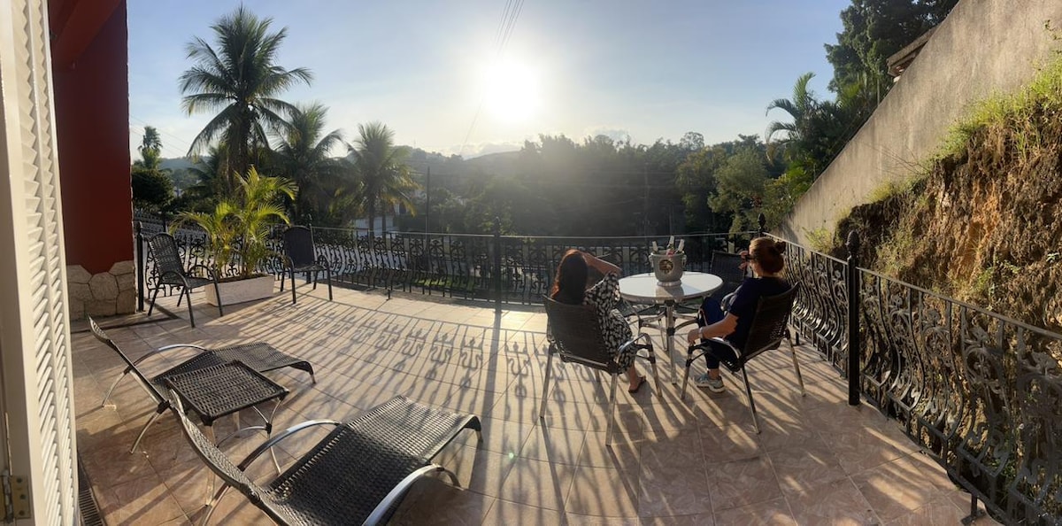 A spacious outdoor terrace is highlighted by comfortable lounge chairs, with a small table situated between them. The setting is enhanced by gentle sunlight casting shadows across the tiled floor, while greenery and distant mountains can be seen beyond the railing.