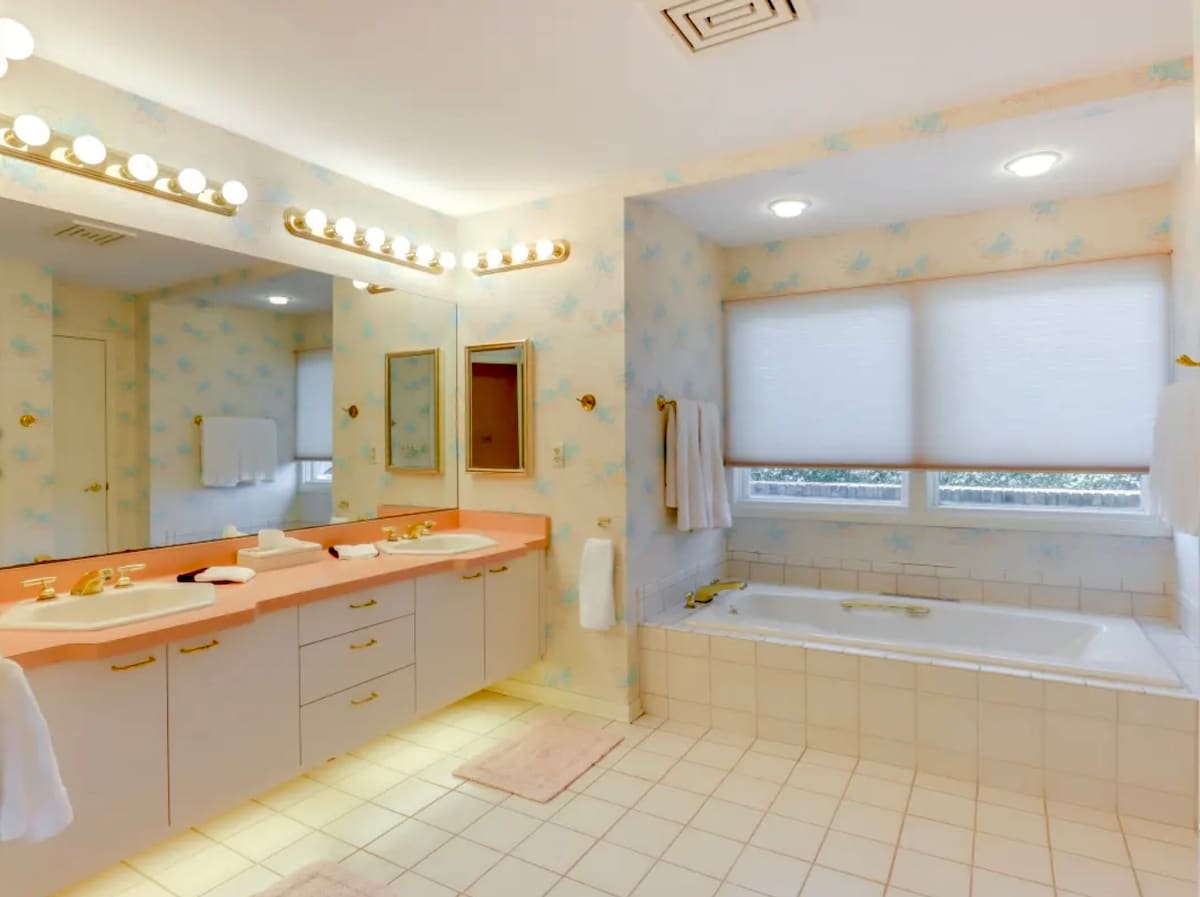 A spacious bathroom includes dual sinks set in a peach-colored countertop, a large soaking tub, and ample storage. Natural light enters through two window coverings. Light-colored tiles adorn the floor, complemented by soft bath mats underfoot.