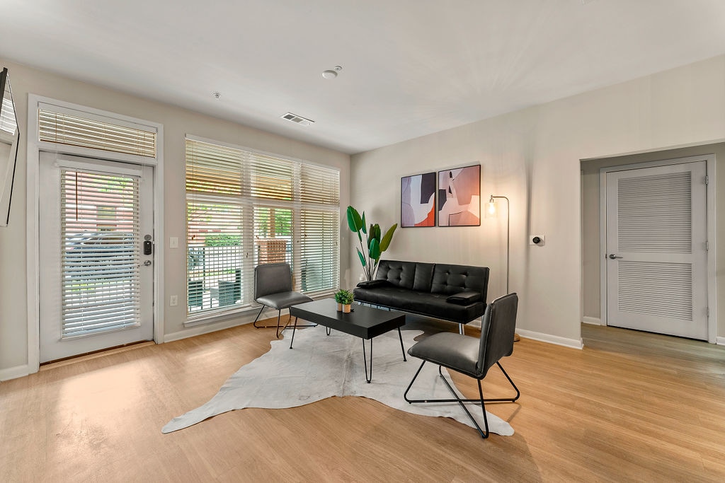 A bright and airy living area features a modern design with large windows allowing natural light to fill the room. A comfortable black sofa is paired with sleek accent chairs, centered around a stylish coffee table atop a light rug. Access to the patio is available through glass doors.